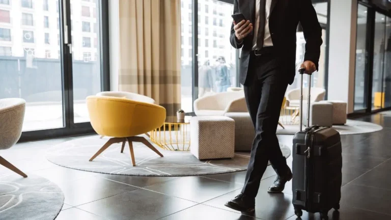 businessman wearing formal black suit holding smartphone and walking with suitcase in hotel lobby